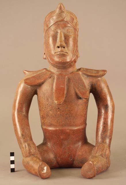 Seated Colima warrior effigy, wearing a helmet and petal-shaped neck decoration.