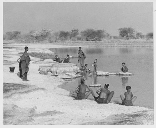 Group of people beside the water; some women are bathing and others are filling ostrich egg shells with water (print is a cropped image)