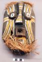 Carved wood mask with yellow and white painted decorations and raffia trim