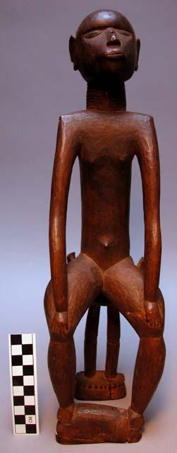 Carved male figure, seated on stool, arms stiffly extended resting on knees.  In