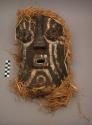 Carved wood mask with black and white painted decorations and raffia trim