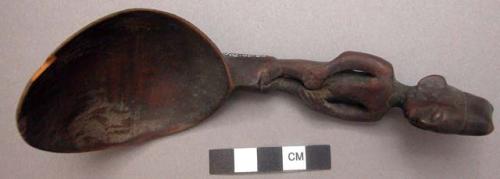 Wooden spoon, handle carved in human effigy: hands resting on flexed knees, sadd