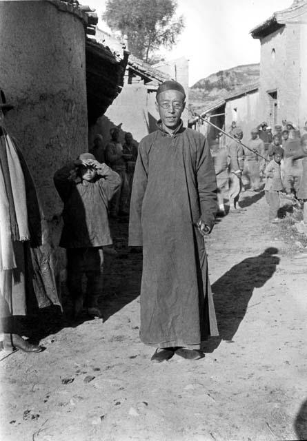 Chinese man in robe and cap, wearing glasses, posed in street