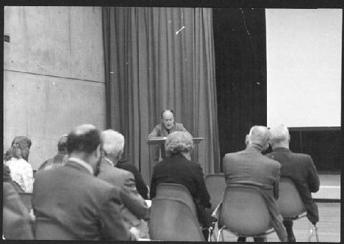 Gordon Willey seated at a desk, speaking to an audience