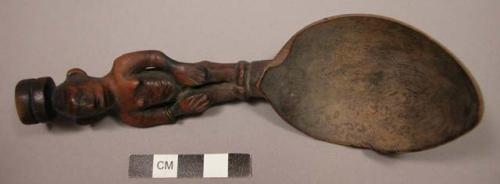 Wooden spoon, handle carved in human effigy: hands resting on flexed knees, coil