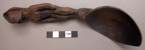 Wooden spoon, handle carved in human effigy: left hand resting on flexed knee, r