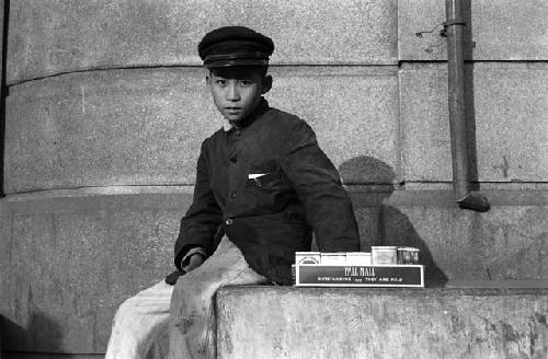 Boy sitting on ledge; selling boxes of Western cigarettes.