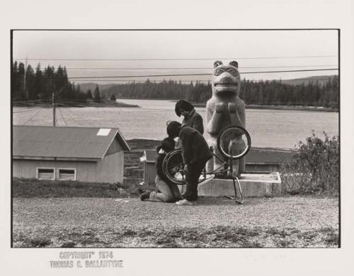 Three children in Hydaburg, Alaska with a bicycle and totem