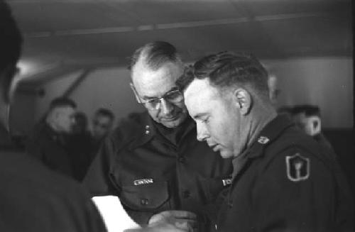 Military man with glasses looks toward man reading paper.