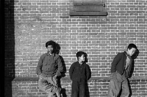 Man and two youths leaning against brick wall.