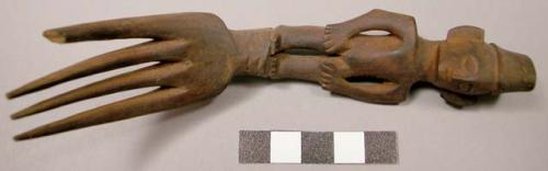 Wooden fork, 4 prongs, handle carved in human effigy: hands resting on flexed kn