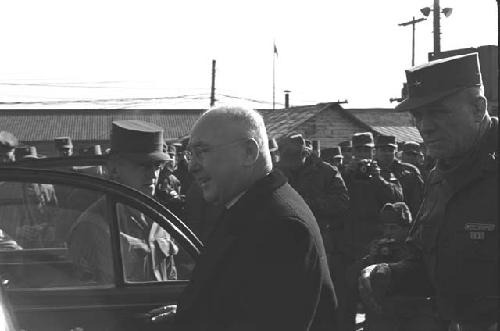 Man with glasses standing next to open car door with military men in crowd.