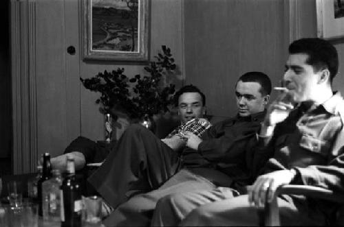 Three men sitting on couch; one smoking; liquor bottles on table.