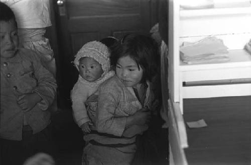 Woman with baby strapped to back; indoors.