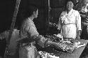 Woman working with slab of meat at a counter;another woman looks on.