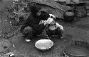 Woman washing face of child outdoors; metal basins on ground.