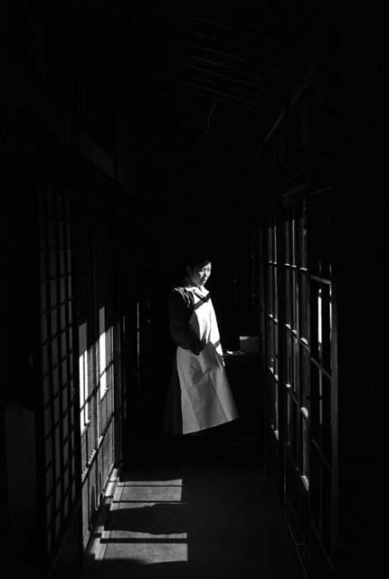 Woman standing in dark corridor, sunlight slants through wooden frames.