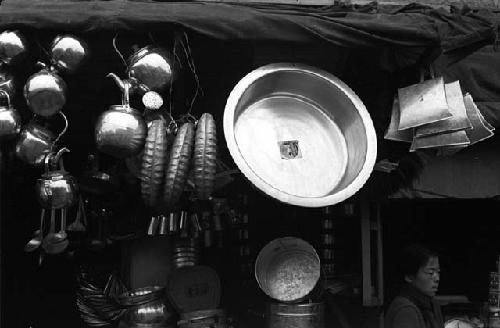 Metal pan, teapots, kitchen accessories hanging from tarp above storefront.