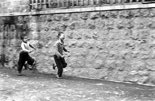 Two boys running down road, beside stone wall.