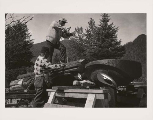 Two men working on a totem pole, one drilling into the back of the pole