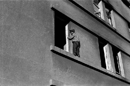 Boy with hat standing on building window ledge.