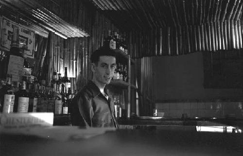 Military man standing in front of bar with liquor bottles.