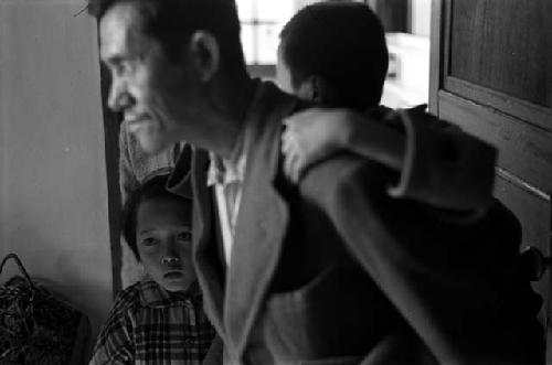 Man with little boy on back, looking left; another child in background.