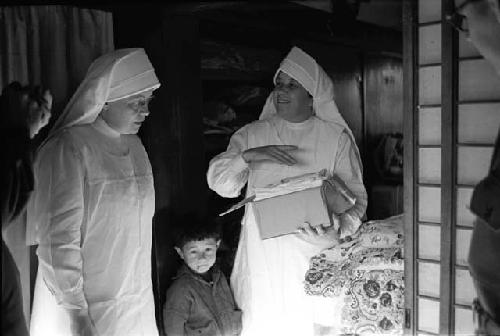 Two nuns speaking; child standing in between; one nun holds box of cloth.