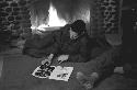 Military man sitting on floor in front of fireplace reading magazine.