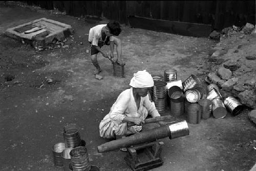 Man working on metal buckets; pile of buckets; child behind him.