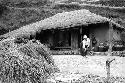 Woman working in front of hut with thatched roof; haystacks in foreground.