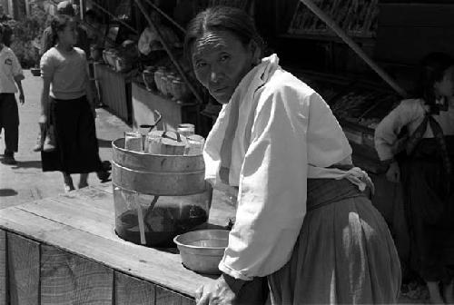 Woman pushing table/cart with some containers down a market street.