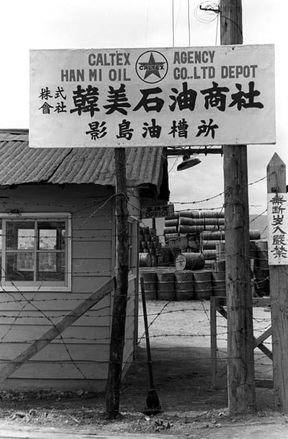 Sign for oil depot nailed to pole; small building; junkyard in background.