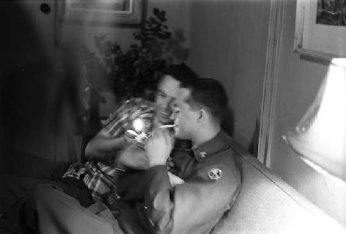Man in plaid shirt lighting the cigarette of the military man.
