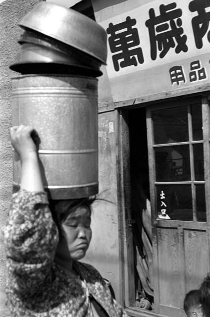 Woman walking by storefront, carrying buckets on her head.