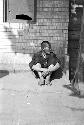 Old man sitting against wall of building with broken window.