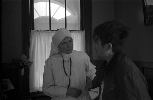 Western nun in white robe talking to man, indoors.