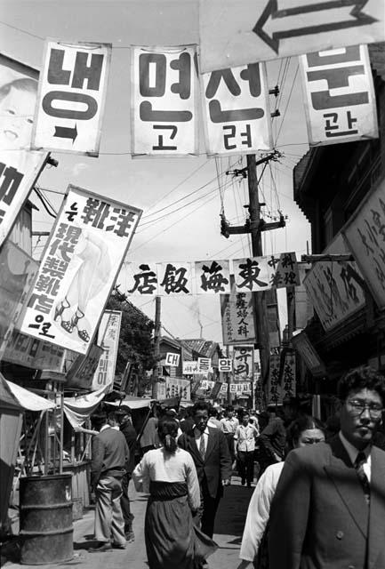 Busy market street, many signs and banners strung above pedestrians