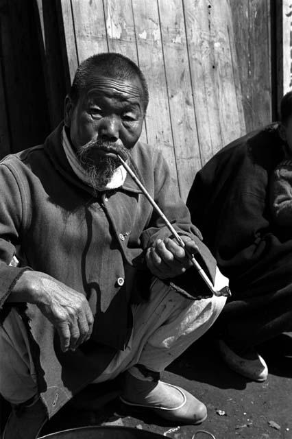 Old man holding a long pipe squatting on ground.