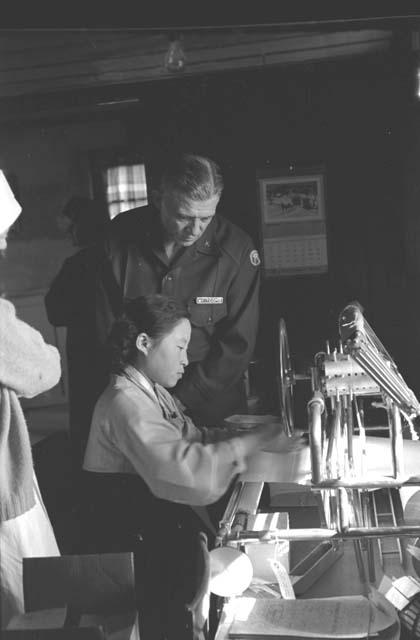 Military Man "Whitcomb" looking as young woman operates machine.