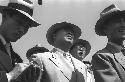 Three men in business suits and hats; more men behind them.