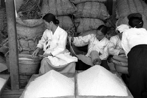 Women sitting and working; piles of grain in front; bags of grain behind.
