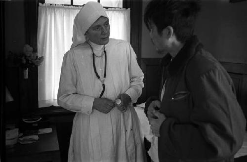 Western nun in white robe talking to man, indoors.