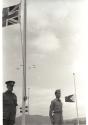 Two soldiers stand near flagpoles at half-mast as Air Force planes pass overhead