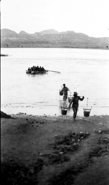 Two men carrying water in buckets on poles to shore, boat on river with people