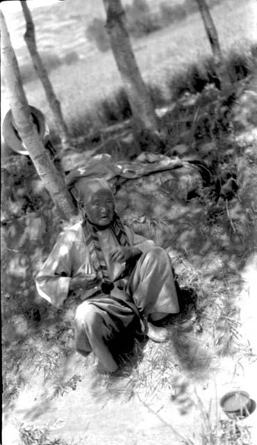 Older woman sitting under trees, bowl in front of her