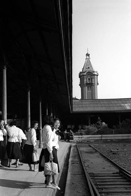 Women in white shirts and dark skirts on platform of train station.