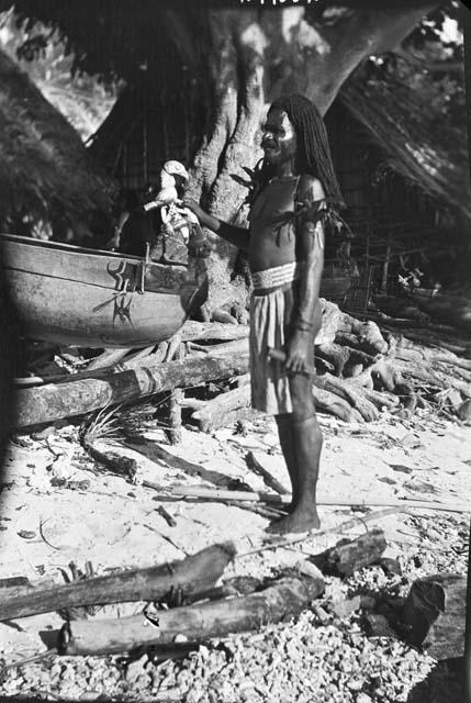 Man standing in front of hut and decorated dugout canoe with bird  sculpture