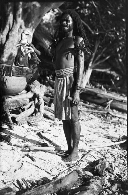 Man standing in front of hut and decorated dugout canoe with bird sculpture
