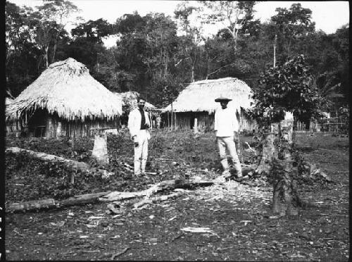 Two men in front of two huts in rural setting